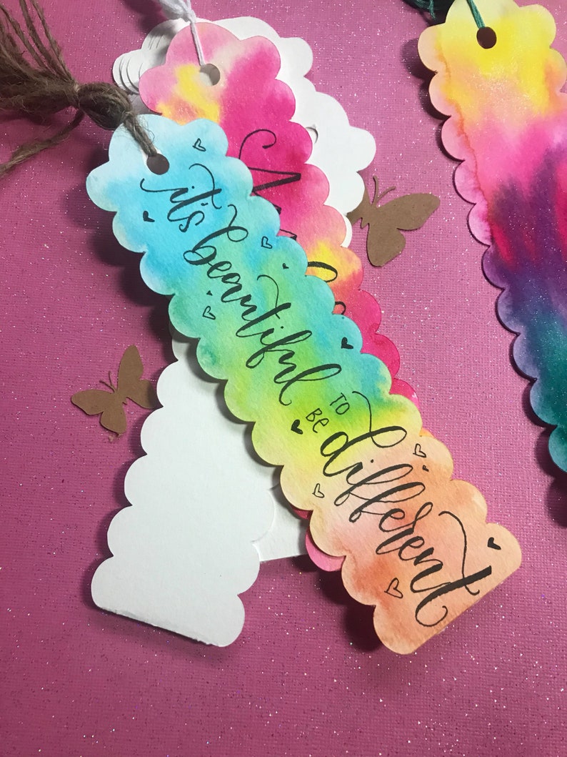 May include: A colorful watercolor bookmark with scalloped edges and the text "It's beautiful to be different". The bookmark has a rainbow gradient of colors, including pink, orange, yellow, green, and blue. There are small hearts and butterflies on the bookmark.