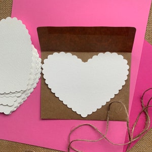May include: A brown envelope with a white heart-shaped card inside. The card has a scalloped edge. The envelope and card are on a pink surface.