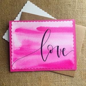 May include: Pink watercolor card with a scalloped edge and the word "love" written in black calligraphy.