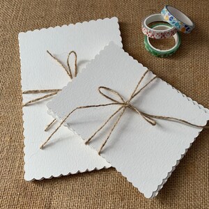 May include: Two white cards with scalloped edges are stacked on top of each other. The cards are tied together with twine and have a rustic, handmade look.
