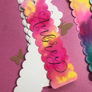 May include: A pink, yellow, and white watercolor bookmark with a scalloped edge. The bookmark has the name "Audrey" written in black calligraphy.