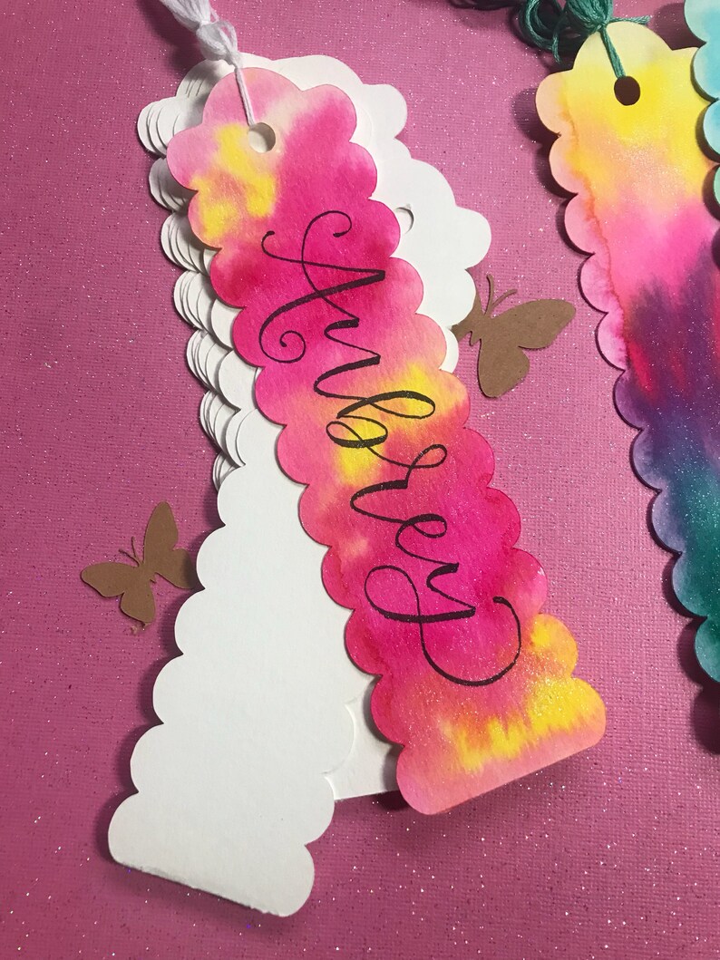 May include: A pink, yellow, and white watercolor bookmark with a scalloped edge. The bookmark has the name "Audrey" written in black calligraphy.