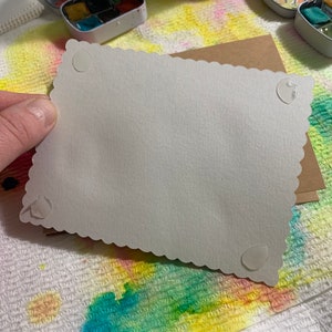 May include: A white card with a scalloped edge and four adhesive dots on the back. The card is blank and ready for writing.