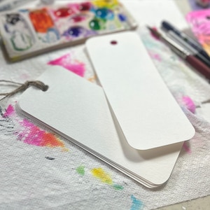 May include: A stack of blank, white rectangular tags with rounded corners and a small hole at the top. One tag has a natural twine string attached. The tags are on a white surface with colorful paint splatters and art supplies in the background.