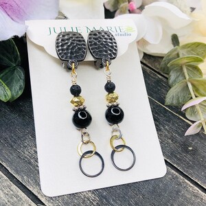 May include: Clip-on earrings with textured, dark gray oval tops. The earrings feature black, gold, and silver beads, with a large black bead and two dangling rings at the bottom. The earrings are displayed on a white card with the text "JULIE MARIE studio".