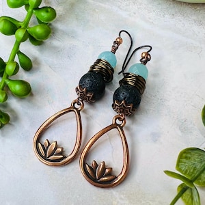 May include: A pair of handcrafted earrings featuring a teardrop-shaped copper frame with a lotus flower design. The earrings have black lava beads, wrapped with copper wire, and topped with a light blue bead. The ear wires are black.
