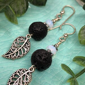 May include: A pair of dangle earrings featuring black lava rock beads, small white beads, and silver-colored leaf charms. The earrings have silver-colored ear wires. The background is a light blue surface with green foliage.