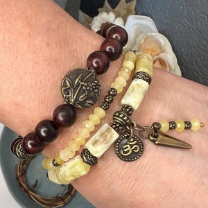 May include: Three beaded bracelets in shades of brown and yellow. One bracelet features large, dark brown beads and a decorative metal accent. Another has yellow beads and rectangular yellow stones. The third bracelet has a charm with the Om symbol.
