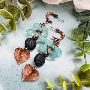 May include: A pair of handcrafted earrings featuring copper-colored metalwork, light blue crystal chunks, and black lava beads. Each earring has a copper leaf charm. The earrings are displayed on a light blue surface with greenery.