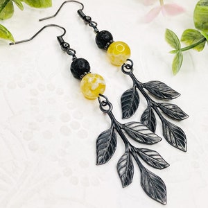 May include: A pair of dangle earrings featuring black metal leaf charms and yellow and black beaded accents. The earrings have a hook closure and are designed with a nature-inspired theme. The earrings are a unique accessory.