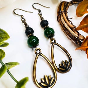 May include: A pair of dangle earrings with a bronze finish. Each earring features a teardrop-shaped pendant with a lotus flower design, a green bead, and a black lava bead. The earrings have bronze-colored ear wires.