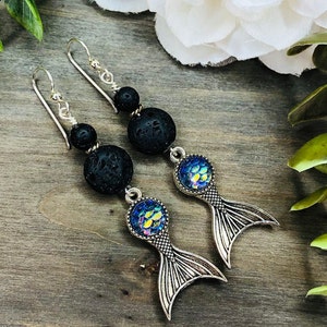 May include: A pair of dangle earrings featuring black lava beads, iridescent blue and yellow scale-like accents, and silver-colored mermaid tail charms. The earrings have silver-colored ear wires and are displayed on a wooden surface.