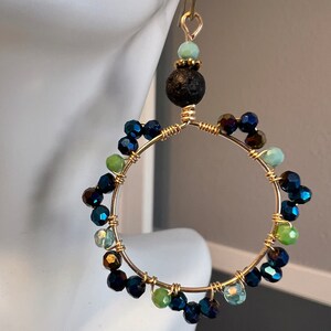 May include: Gold-toned hoop earrings with a cluster of iridescent blue, green, and brown beads. The earrings feature a black lava bead and a small, light green bead at the top. The hoops are suspended from gold-colored ear wires.
