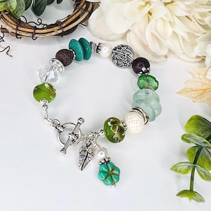 May include: A handmade bracelet with a mix of green, turquoise, brown, and white beads. The bracelet includes clear crystal beads, a silver toggle clasp, and a charm with a leaf and a four-leaf clover. The bracelet is approximately 18 cm in length.