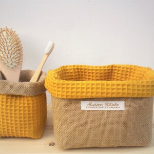 Op de afbeelding: Twee gele opbergmanden van wafelstof met bruine jute zijkanten. De kleinere mand bevat een houten haarborstel en een witte tandenborstel. De grotere mand heeft een label met de tekst "Maison Bibiche Fabrication Française".
