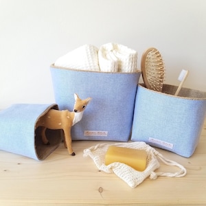 May include: Three light blue fabric storage bins with a natural burlap trim. One bin holds white towels, another a wooden hairbrush and toothbrush. A small deer figurine and a bar of soap in a mesh bag are also present.
