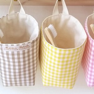 May include: Four hanging fabric storage containers in gray, yellow, and pink gingham patterns. Each container has a white interior and a loop for hanging. Wooden toothbrushes are visible inside some of the containers. The text "Maison Bibiche Fabrication Française" is printed on one.