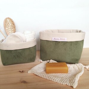 Puede incluir: Dos cestas de tela de pana verde oliva con ribete beige. Una cesta contiene un cepillo de pelo de madera, un cepillo de dientes y otros artículos de aseo. Una pastilla de jabón descansa sobre una bolsa de malla.