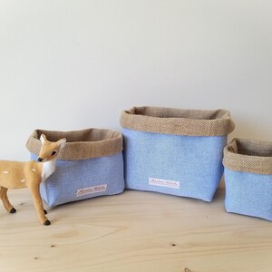 Peut inclure: Trois paniers de rangement en tissu bleu de différentes tailles, avec une bordure en toile de jute. Une figurine de cerf se trouve près des paniers. Chaque panier a une étiquette avec du texte.
