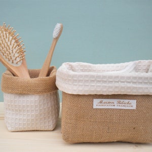 Puede incluir: Dos pequeñas cestas de almacenamiento. Una contiene un cepillo para el pelo y un cepillo de dientes de madera. La otra tiene un forro blanco de tejido de gofre y un exterior de arpillera. La etiqueta dice "Maison Bibiche Fabrication Française".