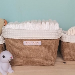 Può includere: Tre cesti di juta marrone con bordatura in tessuto bianco, ognuno riempito con diversi articoli per bambini. Il cestello più grande contiene una pila di pannolini di stoffa bianchi, il cestello centrale contiene una spazzola per bambini bianca e il cestello più piccolo contiene una pila di dischetti di cotone bianchi. Un coniglio di peluche bianco è seduto davanti ai cesti.