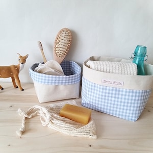 Op de afbeelding: Twee stoffen manden met blauw-witte ruitjesrand, één met een handdoek en een blauwe fles, de andere met een tandenborstel en een haarborstel. Een klein hertje en een stuk zeep in een netzakje zijn ook aanwezig.