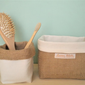 Op de afbeelding: Twee kleine, vierkante opbergmanden van jute met een witte stoffen voering. De mand aan de linkerkant heeft een houten haarborstel en een houten tandenborstel erin. De mand aan de rechterkant heeft een label met de tekst "Maison Bobeche Fabrication Française".
