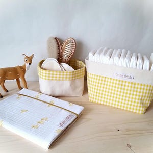 May include: A nursery scene featuring a plush deer, two fabric baskets with yellow gingham trim, and a folded white baby blanket. One basket holds diapers, the other holds cotton pads and brushes. The blanket has a duck pattern.