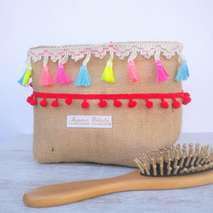 May include: A rectangular burlap bag with colorful tassels and red pom-pom trim. The bag has a white label that reads "Maison Ribiche Fabrication Française". A wooden hairbrush is in the foreground.