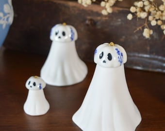 May include: Three white ceramic ghost figurines with blue floral patterns on their heads and small gold accents on top. Each ghost features a painted face with black eyes and a mouth. A black box with a label reading The Little Ghost Company is visible.