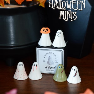 May include: A collection of six small white ghost figurines with various designs, including one wearing an orange pumpkin hat, one with red blood-like splatters, one with red eyes, one olive green, and one with black stitches, displayed with a box.