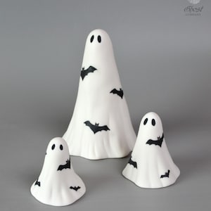 May include: Three white ghost figurines of varying heights, each featuring black oval eyes and small black bat silhouettes printed on the surface. The top right corner displays a logo with a ghost icon and the text The Little Ghost Company.