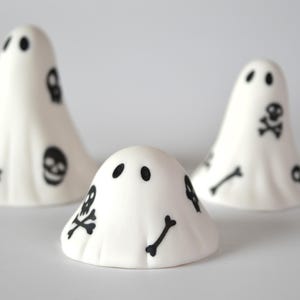May include: Three white ghost figurines of varying heights, each featuring black printed designs of skulls and crossbones. The ghosts have simple black oval eyes and a smooth, rounded shape. A small logo in the top right corner reads The Little Ghost Company.