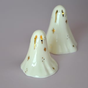 May include: Two white ceramic ghost figurines with smooth, glossy surfaces. Each ghost features gold-painted eye sockets with dripping gold accents and small gold star and dot patterns scattered across their bodies. The figures are rounded.