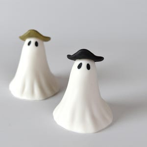 May include: Two small white ghost figurines with smooth, draped bodies and black oval eyes. One ghost wears a metallic gold hat, while the other wears a black hat. The text The Little Ghost Company is visible in the top right corner of the image.
