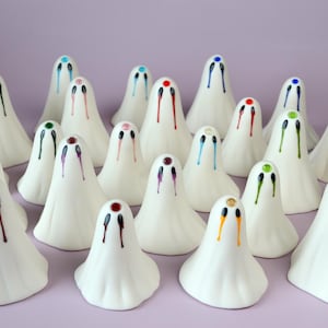 May include: A collection of twenty-two small, white, bell-shaped ghost figurines. Each ghost features two painted eyes with colourful, vertical drips extending downward and a single coloured gem at the top. The logo The Little Ghost Company is visible.