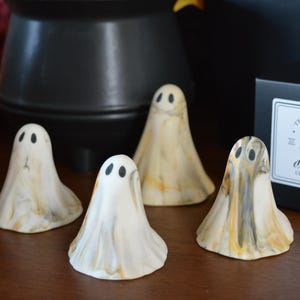 May include: Four small, cone-shaped ghost figurines made of white, marbled material with streaks of grey and gold. Each ghost has two black oval eyes. A black box is present with a white label reading The Little Ghost Company.