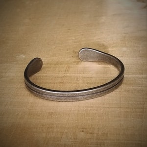 Könnte beinhalten: Ein silberfarbenes Armband mit doppeltem Linienmuster. Das offene Armband besteht aus Metall mit leicht gealtertem Aussehen. Das Armband wird auf einer Holzoberfläche präsentiert.