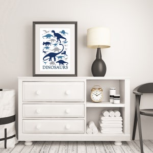 May include: A white dresser with three drawers and a shelf above. The dresser has a framed print of blue and gray dinosaur silhouettes with the word "DINOSAURS" at the bottom. A gold alarm clock, a black lamp with a white shade, and two white cylindrical containers are on the shelf.