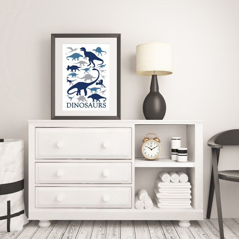 May include: A white dresser with three drawers and a shelf above. The dresser has a framed print of blue and gray dinosaur silhouettes with the word "DINOSAURS" at the bottom. A gold alarm clock, a black lamp with a white shade, and two white cylindrical containers are on the shelf.
