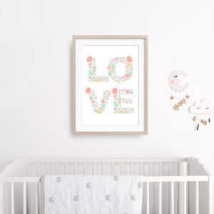 May include: A framed print with the word "LOVE" spelled out in pastel pink and green floral designs. The print is hanging on a white wall above a white crib with a white and gray patterned blanket.