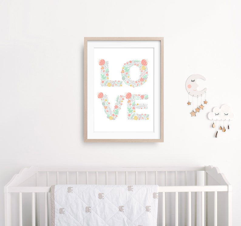 May include: A framed print with the word "LOVE" spelled out in pastel pink and green floral designs. The print is hanging on a white wall above a white crib with a white and gray patterned blanket.