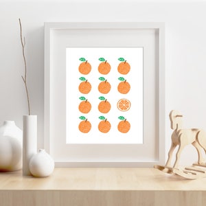 May include: A white framed print with a white background and a pattern of 15 orange citrus fruits with green leaves. One of the fruits is sliced in half and shows the inside of the fruit.