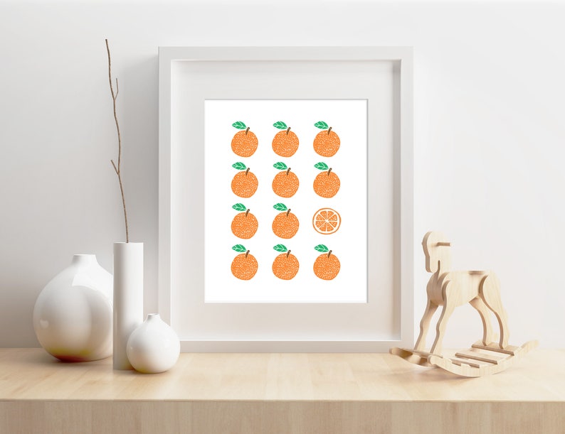 May include: A white framed print with a white background and a pattern of 15 orange citrus fruits with green leaves. One of the fruits is sliced in half and shows the inside of the fruit.