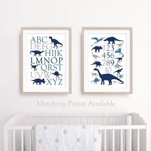 May include: Two framed prints with a blue and gray dinosaur theme. The left print features the alphabet with a dinosaur silhouette for each letter. The right print features the numbers 1-10 with a dinosaur silhouette for each number. The prints are on a white wall above a white crib.