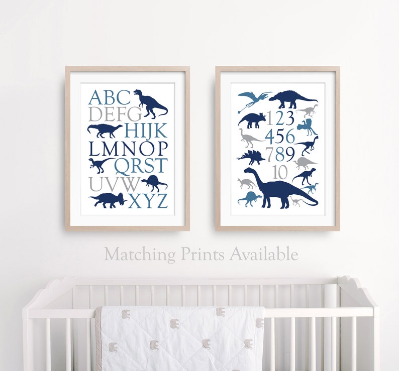 May include: Two framed prints with a blue and gray dinosaur theme. The left print features the alphabet with a dinosaur silhouette for each letter. The right print features the numbers 1-10 with a dinosaur silhouette for each number. The prints are on a white wall above a white crib.