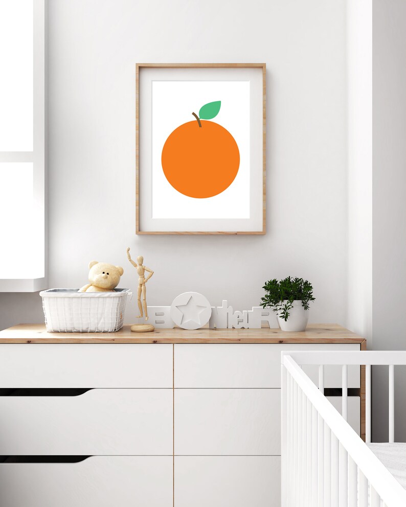 Puede incluir: Una impresi&oacute;n enmarcada de una naranja simple con una hoja verde sobre un fondo blanco. La impresi&oacute;n est&aacute; colgada en una pared blanca sobre una c&oacute;moda blanca con una tapa de madera. Hay un oso de peluche, una mu&ntilde;eca de madera, una estrella blanca y un letrero de madera que dice "Hola" en la c&oacute;moda.