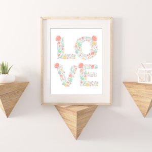 May include: A framed print with the word "LOVE" spelled out in floral letters. The letters are made up of pink, green, and yellow flowers and leaves. The print is hanging on a white wall with two wooden shelves on either side.