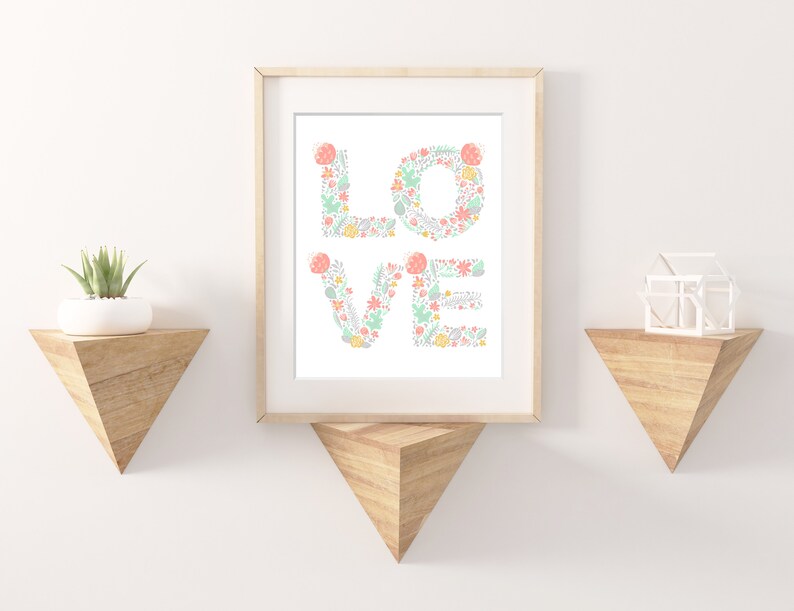 May include: A framed print with the word "LOVE" spelled out in floral letters. The letters are made up of pink, green, and yellow flowers and leaves. The print is hanging on a white wall with two wooden shelves on either side.