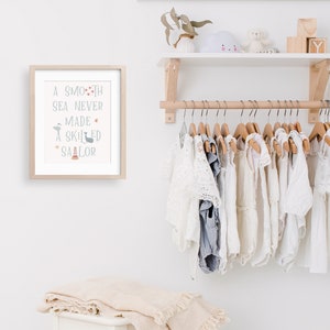 May include: A framed print with the text "A smooth sea never made a skilled sailor" with a nautical theme. The print is hanging on a white wall above a wooden shelf with a white surface. The shelf is holding a white cloud-shaped stuffed toy, a small wooden box, and a wooden block with the letter "Y" on it. Below the shelf is a wooden rod with a row of white clothing hanging on wooden hangers.